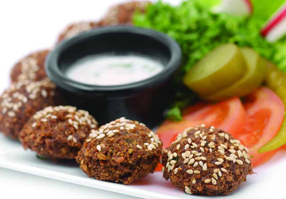 Golden falafel balls topped with sesame seeds served with fresh vegetables and creamy dipping sauce on a white plate.